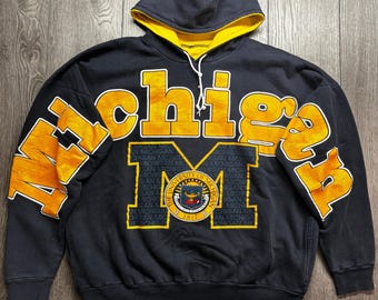May include: A navy blue hooded sweatshirt featuring the word Michigan in large yellow letters across the chest and sleeves. A large yellow letter M is centred with the University of Michigan seal and the text 1817 printed inside the emblem.