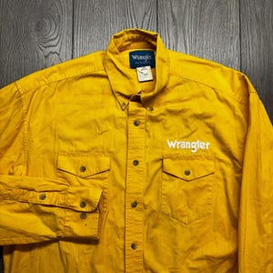 Puede incluir: Camisa Wrangler amarilla mostaza con botones. La camisa tiene dos bolsillos en el pecho, cuello abotonado y el logotipo de Wrangler bordado en blanco en la parte delantera. Mangas largas con puños abotonados.