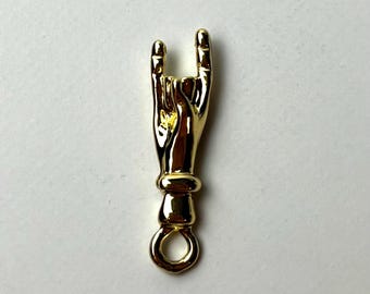 Könnte beinhalten: Ein goldfarbener Anhänger in Form einer Hand, die die Geste „Rock on“ macht. Der Anhänger hat unten eine Schlaufe zum Befestigen an einer Halskette oder einem Armband. Die Hand ist mit sichtbaren Fingern und einem Armband detailliert.