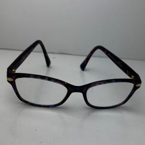 May include: Spectacles with a marbled pattern in shades of purple, blue, and black. The frames have a cat-eye shape with gold accents at the hinges. The lenses are clear.