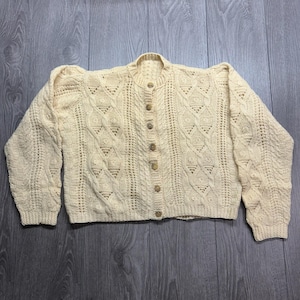May include: A pale yellow long-sleeve cardigan featuring a complex cable knit pattern with vertical columns of diamond shapes and textured bobbles. The garment has a round neckline, ribbed cuffs, and a front button closure with seven matching buttons.