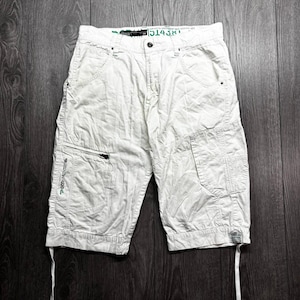 May include: Off-white cargo shorts featuring a button and zip fly closure, multiple pockets including a zipped side pocket with green text, and a small rectangular patch on the lower leg. The waistband interior displays the number 34.
