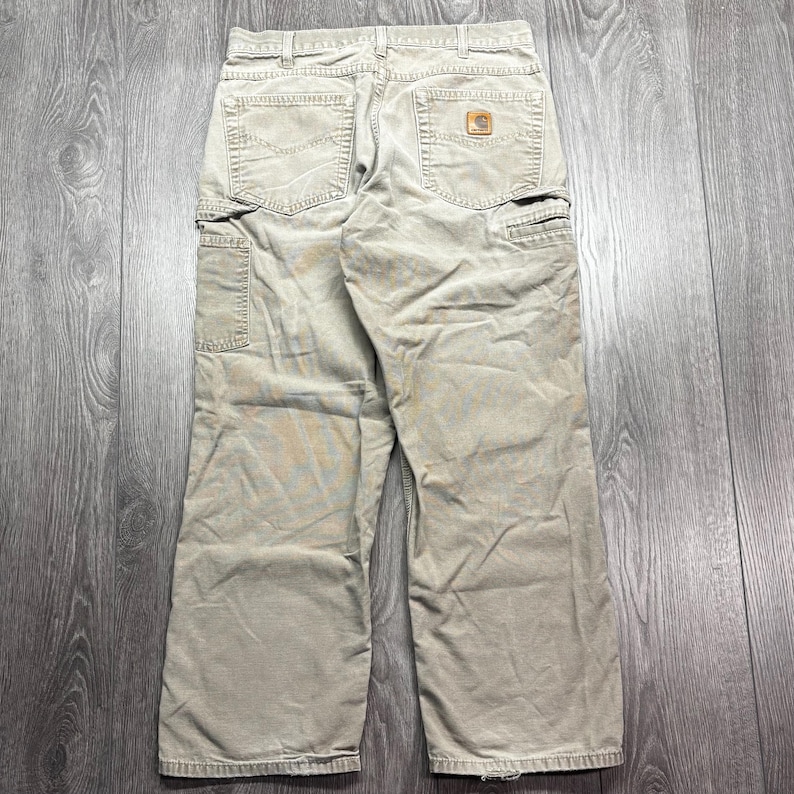 Puede incluir: Pantalones de trabajo color caqui con m&uacute;ltiples bolsillos, incluyendo dos bolsillos traseros y un bolsillo lateral. Los pantalones tienen un dise&ntilde;o de pierna recta y un peque&ntilde;o parche cuadrado con el logo en el bolsillo trasero. La tela parece duradera y ligeramente desgastada.