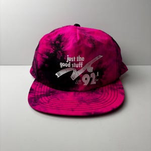 Puede incluir: Gorra de béisbol rosa y negra con teñido anudado con el texto "just the good stuff" y "92.1 fm" en blanco. La gorra tiene una visera plana y un botón negro en la parte superior.