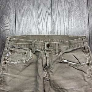 Puede incluir: Primer plano de unos pantalones vaqueros de color caqui con cierre de bot&oacute;n y cremallera. Los pantalones tienen bolsillos delanteros y trabillas para el cintur&oacute;n. La tela parece desgastada, con algunos deshilachados alrededor de los bolsillos.