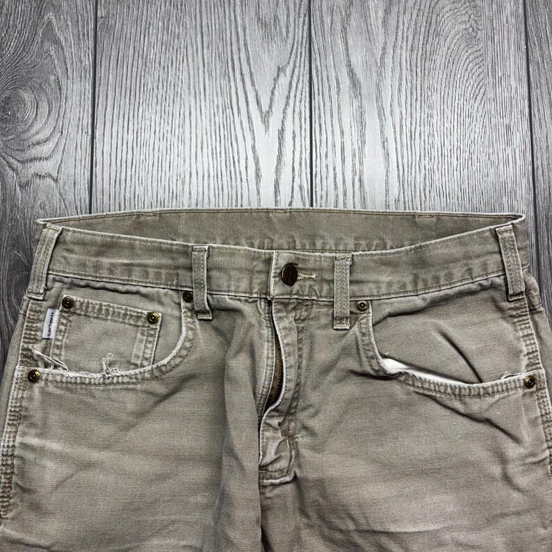 Puede incluir: Primer plano de unos pantalones vaqueros de color caqui con cierre de bot&oacute;n y cremallera. Los pantalones tienen bolsillos delanteros y trabillas para el cintur&oacute;n. La tela parece desgastada, con algunos deshilachados alrededor de los bolsillos.