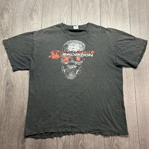 Puede incluir: Una camiseta gris carbón de manga corta con un gráfico de un cráneo robótico metálico con ojos rojos brillantes. El texto "TERMINATOR SALVATION" está impreso en blanco y rojo en el centro del pecho, posicionado sobre el cráneo.