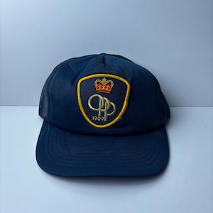 Puede incluir: Gorra de camionero azul marino con visera curva y parte trasera de malla. La parte delantera presenta un parche con borde dorado, una corona roja y las letras "QP" doradas, con el año "1909" debajo. Un accesorio clásico.