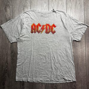 May include: A heather gray short-sleeve t-shirt featuring the band name AC/DC printed in a bold, red and yellow gradient font across the chest. The shirt has a classic crew neckline and a standard fit, showcasing a simple and casual design.