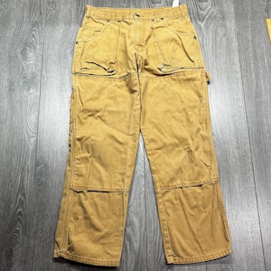 Puede incluir: Pantalones de trabajo color canela con múltiples bolsillos y rodillas reforzadas. Los pantalones tienen un diseño clásico de ropa de trabajo con cierre de botón y cremallera. La tela parece ser una mezcla de algodón duradera.