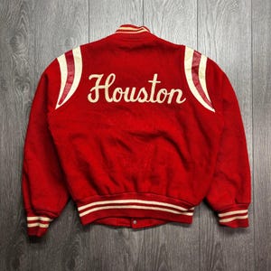May include: A red varsity jacket featuring the word Houston in white script lettering across the back. The jacket has white and red leather shoulder accents, a ribbed collar, and striped ribbed cuffs and hem in red and white.