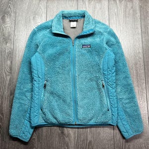 May include: A turquoise fleece jacket with a full zipper closure. The jacket features a stand-up collar, two zippered pockets, and a small logo patch on the chest. The jacket is made of a soft, textured material.