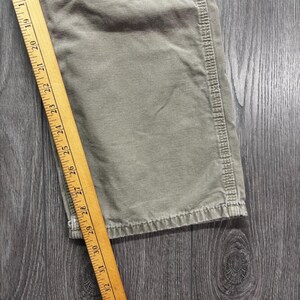 Puede incluir: Primer plano de tela verde oliva con una regla de madera que mide de 48 a 81 cm. La tela parece ser una secci&oacute;n de pantal&oacute;n o prenda similar, con costuras visibles en el borde.