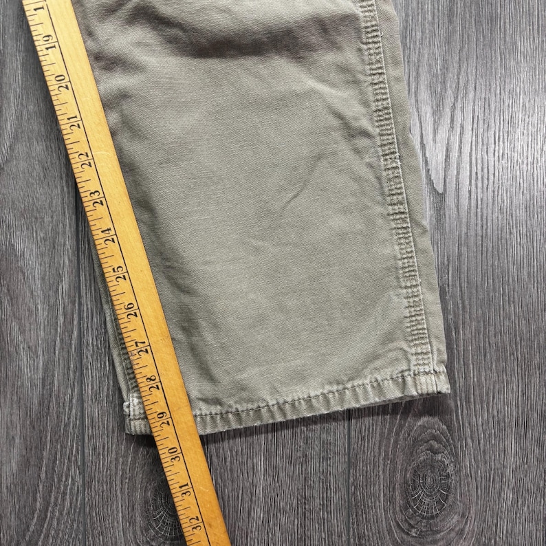 Puede incluir: Primer plano de tela verde oliva con una regla de madera que mide de 48 a 81 cm. La tela parece ser una secci&oacute;n de pantal&oacute;n o prenda similar, con costuras visibles en el borde.