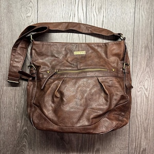 May include: A brown leather shoulder bag with a long adjustable strap. The bag has a front zipper pocket and a small metal tag that reads "ROXY". The bag is made of a textured leather material.