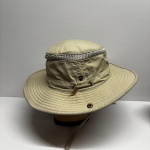 May include: A beige wide-brimmed hat featuring a mesh ventilation band around the crown, a drawstring cord with a small tag labeled Tilley, and a metal snap button on the side of the brim. The hat has a structured top and a downward sloping brim.