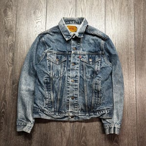 May include: A faded blue denim jacket with a button-up front, two chest pockets, and a classic collar. The jacket has a worn appearance and a small red tag on the chest pocket. The jacket is displayed on a wooden surface.