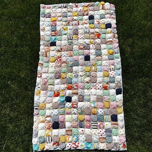 Pode incluir: Uma colcha retangular construída a partir de numerosos pequenos quadrados de tecido acolchoados em uma variedade de cores e padrões, incluindo designs florais, xadrez, listrados e sólidos, dispostos em uma grade com uma borda azul clara.
