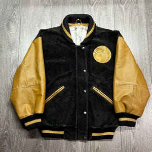 May include: Black and tan varsity jacket with a round patch on the chest. The jacket features tan leather sleeves, a black body, and gold and black striped trim on the collar, cuffs, and hem. The jacket has a button-up front and two front pockets.