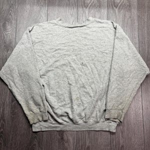 Puede incluir: Una sudadera gris jaspeada con cuello redondo y mangas largas. Los pu&ntilde;os y el dobladillo muestran signos de desgaste. La prenda est&aacute; tendida sobre una superficie de madera.