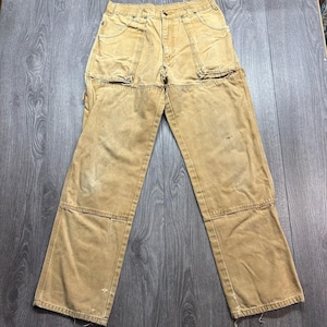 Puede incluir: Pantalones de trabajo color canela con construcción de algodón resistente, rodillas reforzadas de doble capa, múltiples bolsillos utilitarios, cierre de botones y trabillas para cinturón. La tela muestra desgaste con manchas y deshilachados.