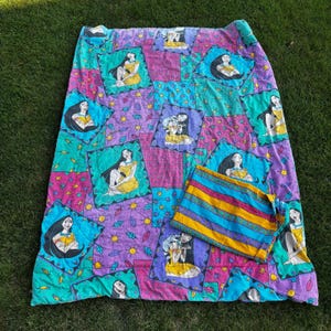 May include: A quilted blanket featuring a repeating pattern of Pocahontas in diamond-shaped frames against a purple and teal background with small leaf and sun motifs. A folded section displays horizontal stripes of yellow, blue, and purple.