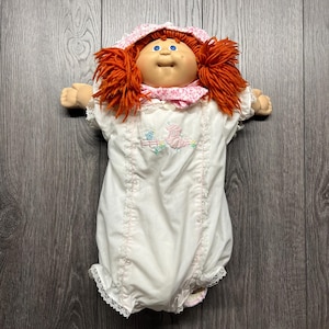 Puede incluir: Muñeca Cabbage Patch Kid vintage con cabello de hilo naranja y ojos azules. La muñeca viste un pelele blanco con ribete floral rosa y un gorro a juego. El pelele presenta un diseño bordado de un cerdo y una valla.