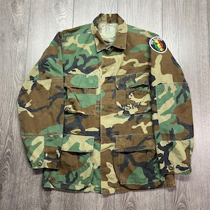 Może przedstawiać: Kurtka w kamuflażu z naszywką Boba Marleya na lewym rękawie i haftem "USMC" po prawej stronie. Kurtka ma wiele kieszeni i zapięcie na guziki.