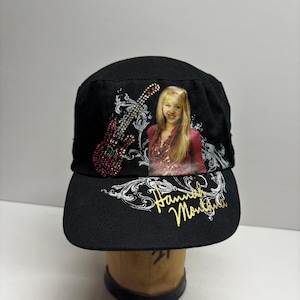 Peut inclure: Une casquette de style militaire noire ornée d'un graphique représentant une personne aux cheveux blonds, une guitare rouge décorée de strass rouges, des volutes blanches et le texte Hannah Montana en écriture cursive dorée.
