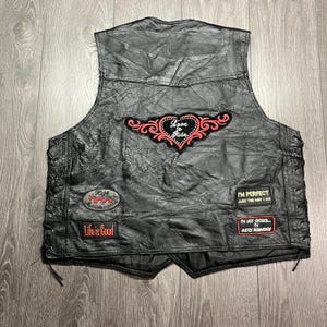 May include: Black leather vest with various patches. Patches feature text such as "Love to Ride", "High Maintenance", "Life is Good", "I'm Perfect Just the Way I Am", and "I'm Not Stored I'm Justly Rewinded". The vest has a lace-up design on the sides.