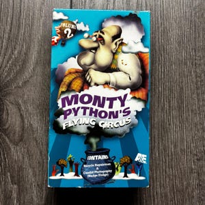 Może przedstawiać: Okładka kasety VHS Monty Python's Flying Circus Volume 2. Niebieska okładka przedstawia postać z kreskówki z wyłupiastymi oczami, balon na ogrzane powietrze z napisem Volume 2 oraz czarny kocioł wydobywający dym z tekstem o zawartości skeczy.