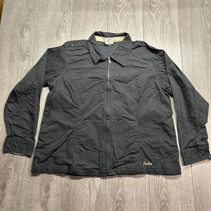 Peut inclure: Une veste gris anthracite à manches longues avec une fermeture éclair intégrale sur le devant, un col pointu et des poignets boutonnés. Le vêtement présente un petit logo Dickies brodé sur le bas et une doublure de col claire avec une étiquette.