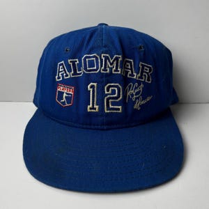 May include: A blue baseball cap with the name "ALOMAR" embroidered in white, along with the number "12" and a signature. The cap also features a "PLAYERS" logo. The hat has a curved brim.