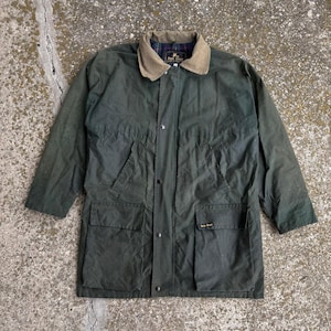 May include: A dark olive green waxed cotton jacket featuring a tan corduroy collar, a front snap-button closure, two large lower flap pockets, and two slanted chest pockets. The interior is lined with a dark blue, red, and green plaid fabric.