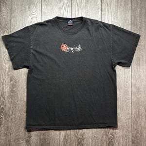 May include: A dark grey t-shirt with a crew neck and short sleeves. The shirt features a graphic in the centre with the text "Rob Zombie" and a red and white design. The shirt has a vintage, worn appearance.