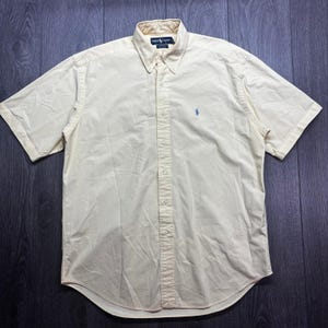 Può includere: Camicia a maniche corte giallo chiaro con colletto button-down. Presenta un piccolo logo blu sul petto. La camicia è realizzata in tessuto leggero e ha una chiusura con bottoni.