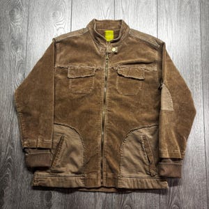 May include: Brown corduroy jacket with a zip-up front, two chest pockets, and two lower pockets. The jacket has a stand-up collar and ribbed cuffs. The fabric has a distressed look.