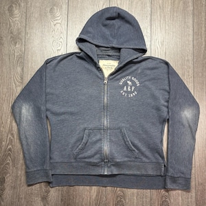 May include: A blue zip-up hoodie with a hood and a kangaroo pocket. The hoodie has the text "QUALITY GOODS A&F EST. 1892" printed on the front. The hoodie is made of a soft, comfortable material.