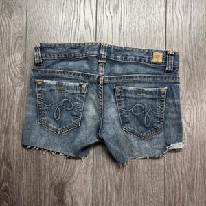 Könnte beinhalten: Blaue Jeansshorts in mittlerer Waschung mit ausgefransten Säumen und zwei Gesäßtaschen mit Ziernähten. Der Bund ist mit Gürtelschlaufen und einem kleinen, rechteckigen, lederartigen Aufnäher auf der rechten Rückseite mit Text versehen.