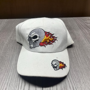 May include: White baseball cap with an embroidered skull and flames design. The skull is gray with black eyes and a red mouth. The flames are red, yellow, and orange.