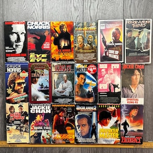 Puede incluir: Una colección de cintas VHS vintage con películas de acción y artes marciales. Los títulos incluyen "The Running Man", "Hellbound", "Code of Silence", "Drunken Master" y otros. Las cintas están dispuestas sobre una superficie de madera.