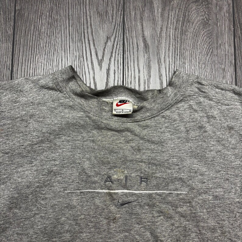 Puede incluir: Una sudadera gris jaspeado con cuello redondo y el logo de Nike en el cuello. La palabra "AIR" est&aacute; bordada en blanco en el pecho, con un peque&ntilde;o logo de Nike swoosh debajo. La sudadera est&aacute; hecha de un material suave y c&oacute;modo.