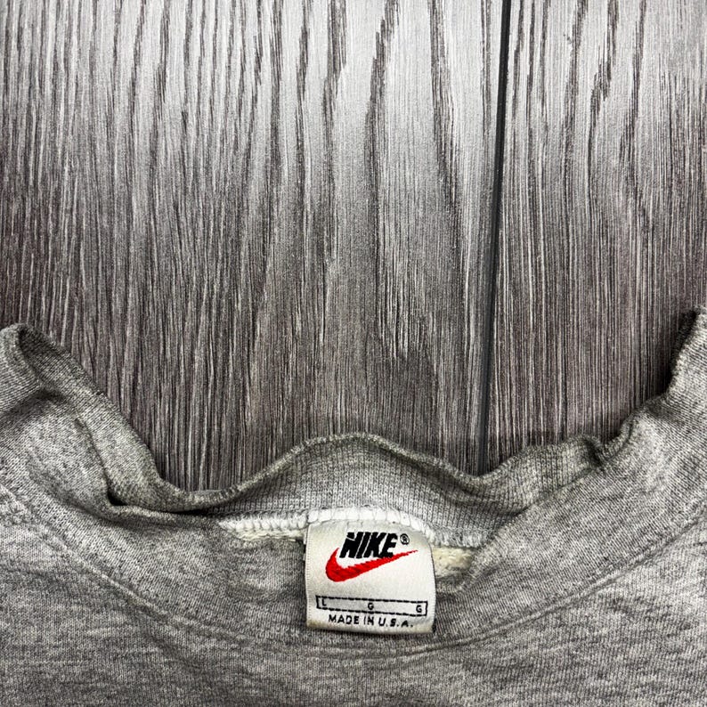 Puede incluir: Primer plano del cuello de una sudadera Nike gris. La etiqueta presenta el logotipo de Nike en rojo y blanco, con el texto "MADE IN U.S.A." La sudadera est&aacute; hecha de un material suave y texturizado.