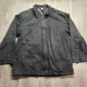 Puede incluir: Una chaqueta negra de manga larga con cremallera frontal completa, cuello vuelto y dos bolsillos delanteros inclinados. El interior cuenta con un forro gris y una etiqueta de la marca Tommy Hilfiger visible en el centro del cuello.