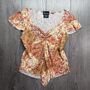 May include: A short-sleeve top with a V-neckline and a gathered centre front detail. The garment features a patchwork-style floral print in shades of orange, red, tan, and cream. A black label at the inner neckline displays the text No Boundaries.