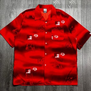 Op de afbeelding: Rood overhemd met korte mouwen en een zwart-rood gestreept patroon. Het shirt heeft wit omlijste illustraties van een persoon en een hersenen. Het shirt heeft een kraag en een borstzak.