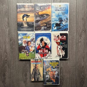 Op de afbeelding: Een verzameling VHS-banden met familiefilms zoals "Free Willy", "Heidi" en "101 Dalmatiërs". De banden hebben kleurrijke covers met filmtitels en afbeeldingen, waaronder walvissen, honden en mensen. De banden zijn gerangschikt op een houten oppervlak.