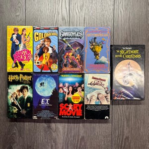 Op de afbeelding: Een verzameling VHS-banden met filmtitels als "Austin Powers", "Goldmember", "Gargoyles" en "The Nightmare Before Christmas". De banden hebben kleurrijke covers met filmkunst en tekst, gerangschikt op een houten oppervlak.