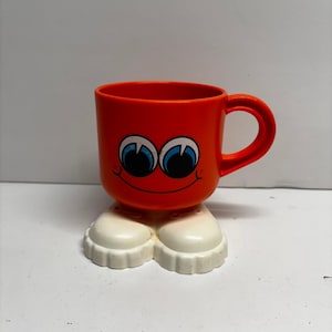 May include: A bright orange mug featuring a cartoon face with large blue and black eyes and a wide smiling mouth. The mug is supported by a base shaped like a pair of white shoes with thick, textured soles, providing a stable foundation for the cup.
