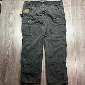 May include: Dark gray Wrangler cargo pants with multiple pockets and reinforced knees. The pants have a faded, worn appearance and a brown patch on the left pocket. The Wrangler logo is visible on the back.